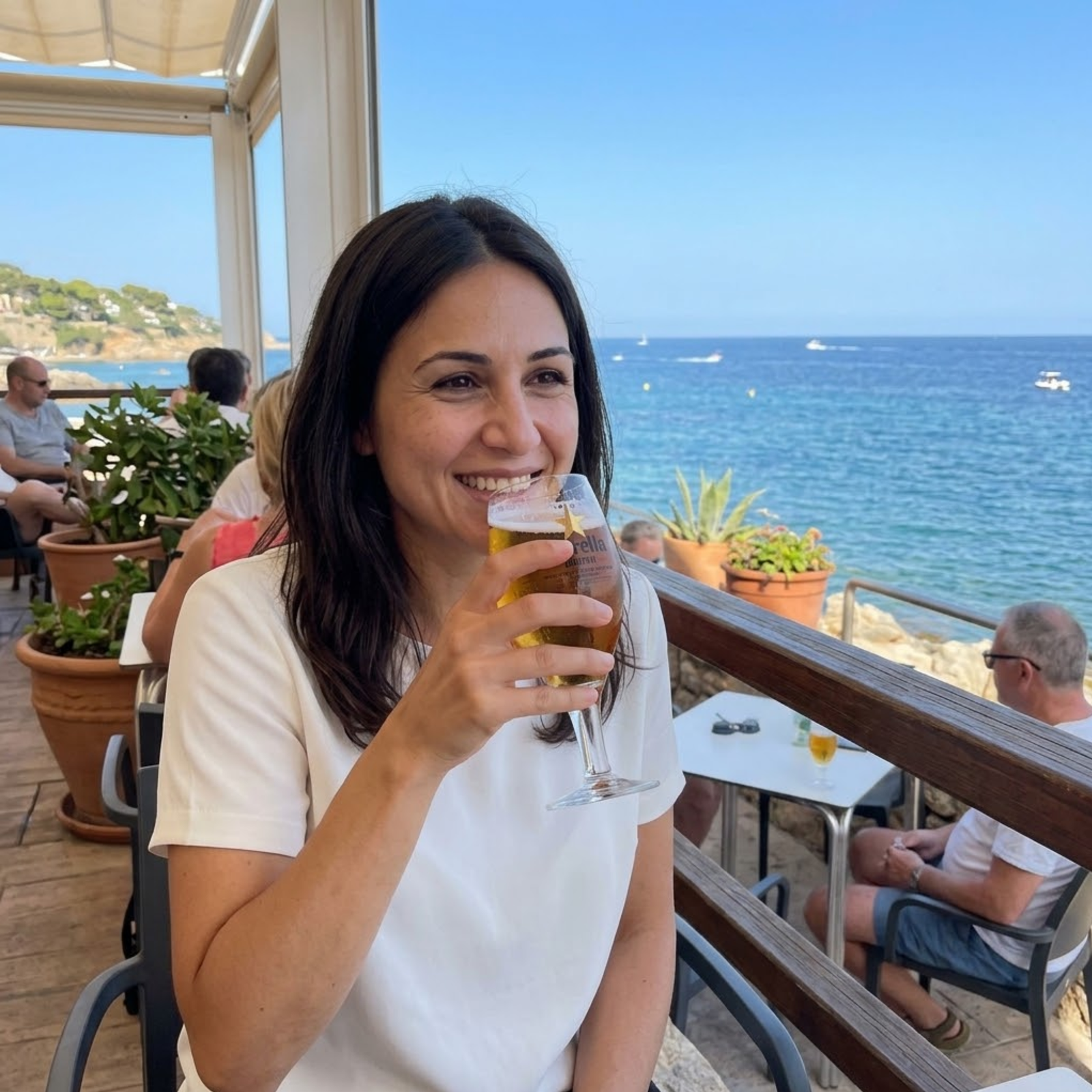 Cristina sonríe mientras toma una cerveza en una terraza junto al mar, con cielo despejado y varias embarcaciones al fondo.